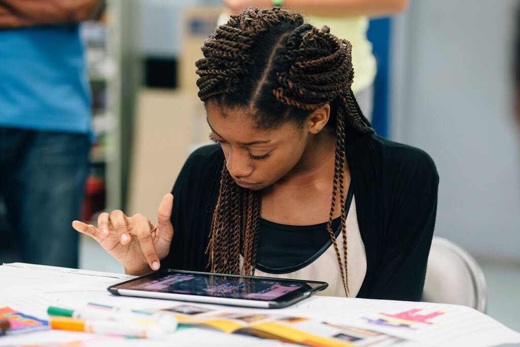 A teen sitting at a table of markers and papers using a digital tablet