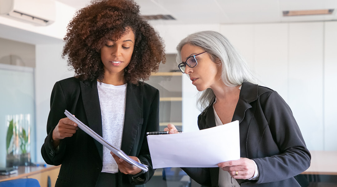 Two people in business suits discuss reports they are holding.