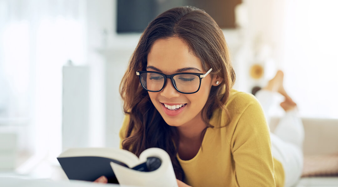 Person in yellow shirt and black glasses smiles while reading a book.