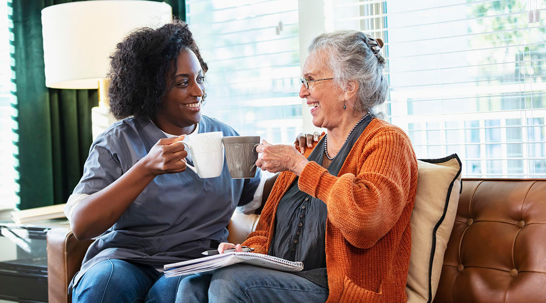 A senior in their 80s sitting on a couch, having coffee and conversing with a younger adult.