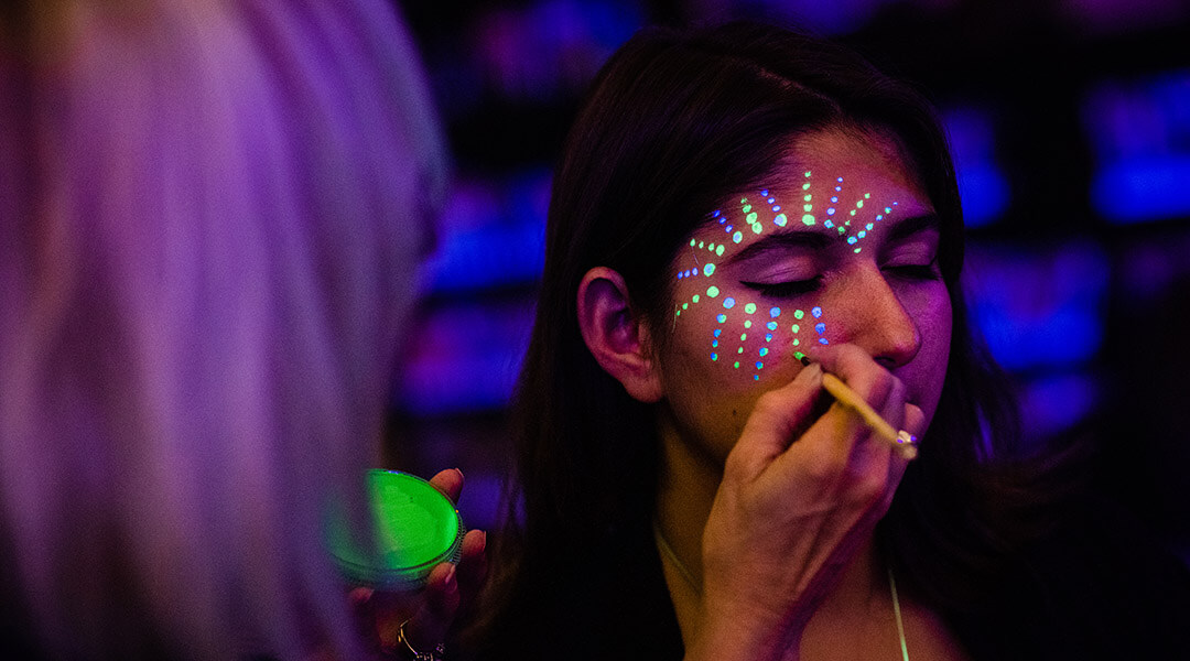 A teenager gets their face painted with neon face paint.