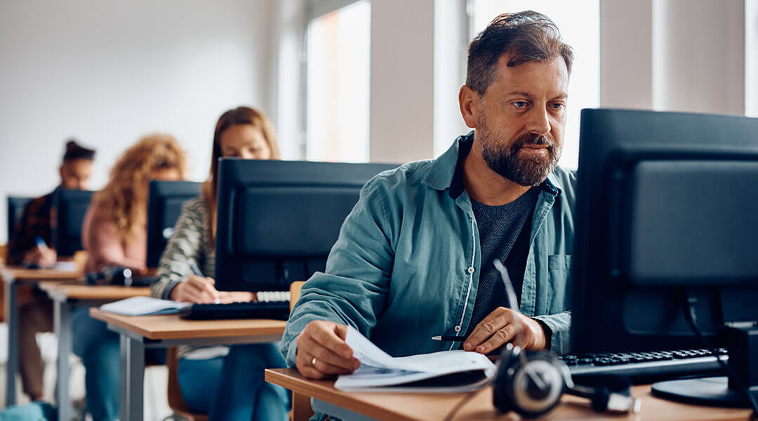 A middle aged person sitting at a computer with a workbook with others at their own computers in the background.