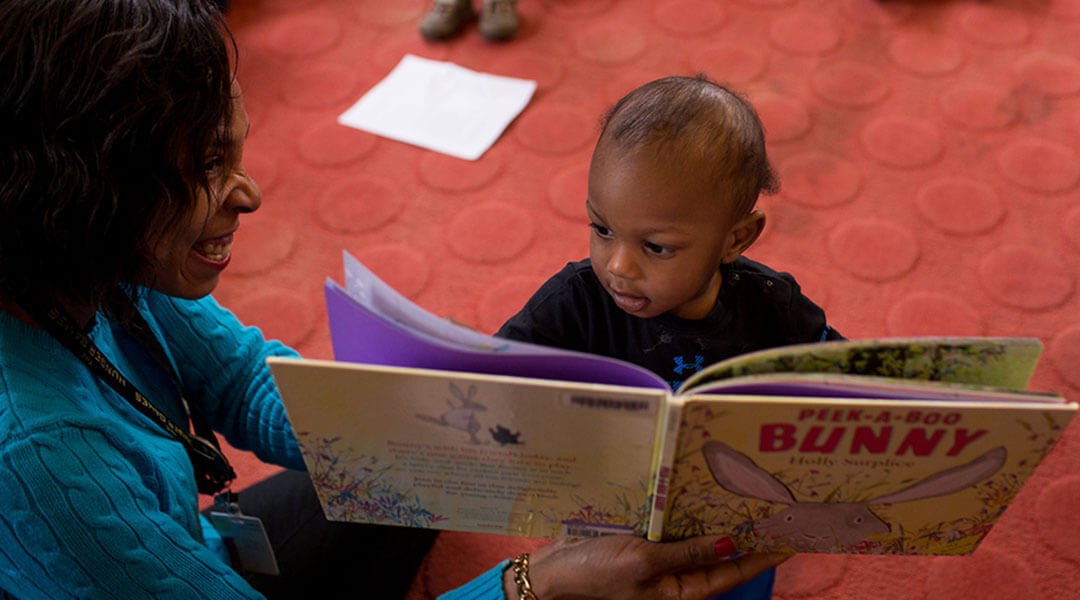 A librarian holds up a picture book during storytime and a baby stands in front of it to investigate.