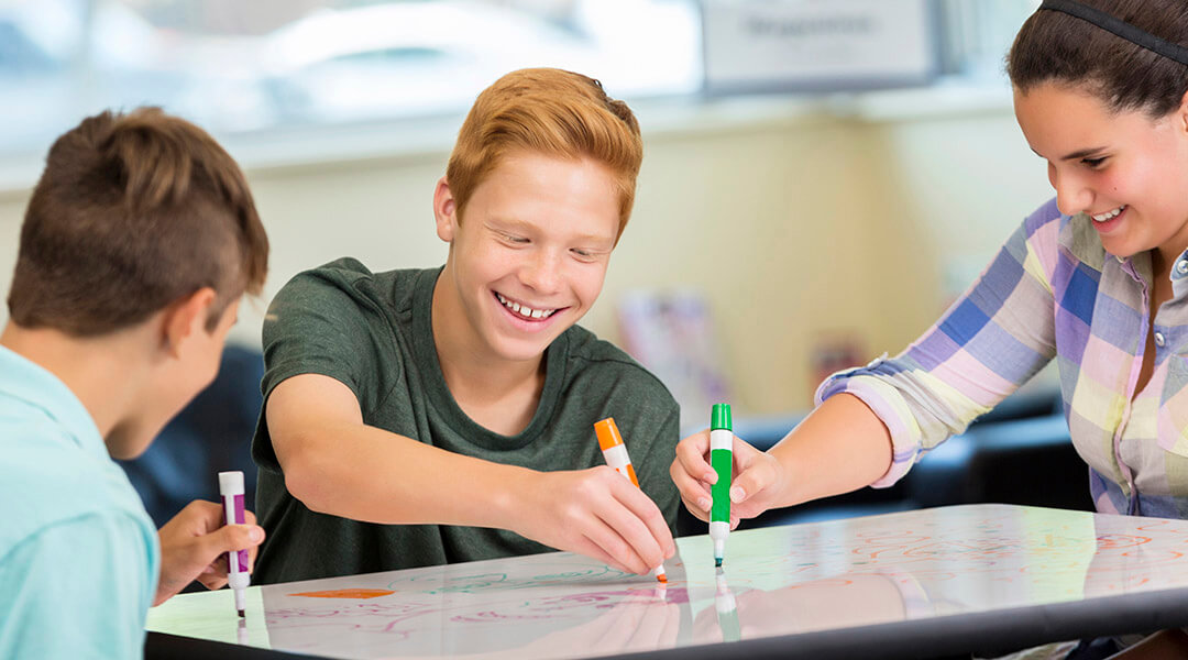 Three teenagers write on a flat whiteboard that has been set up between them.