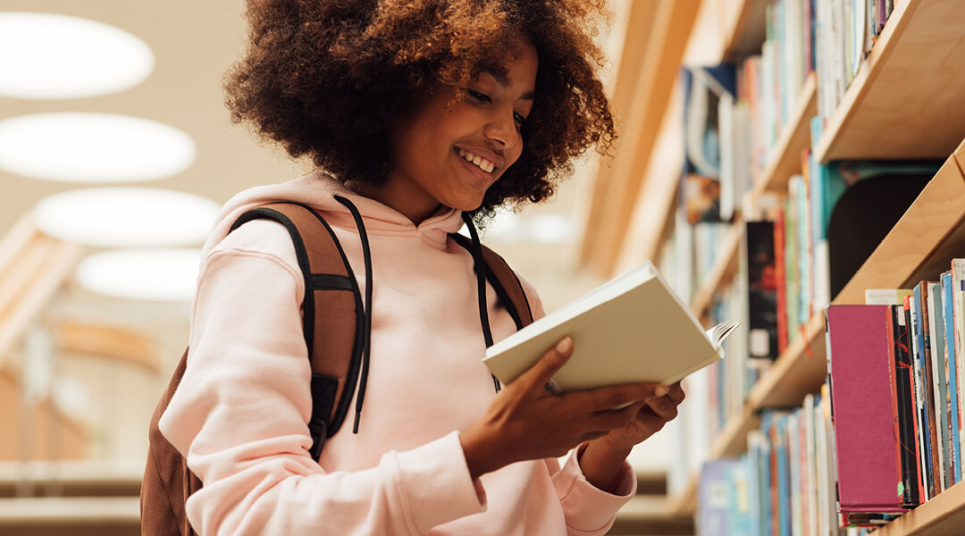 Teenager smiling while reading a book in front of a library bookshelf.