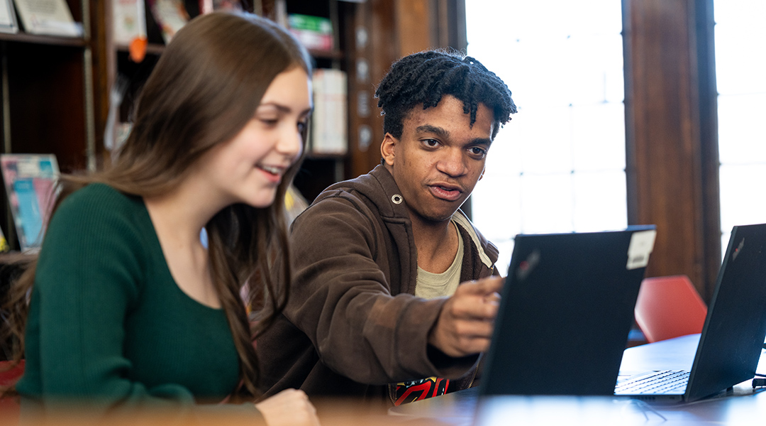 Two young adults compare laptop screens in the library.