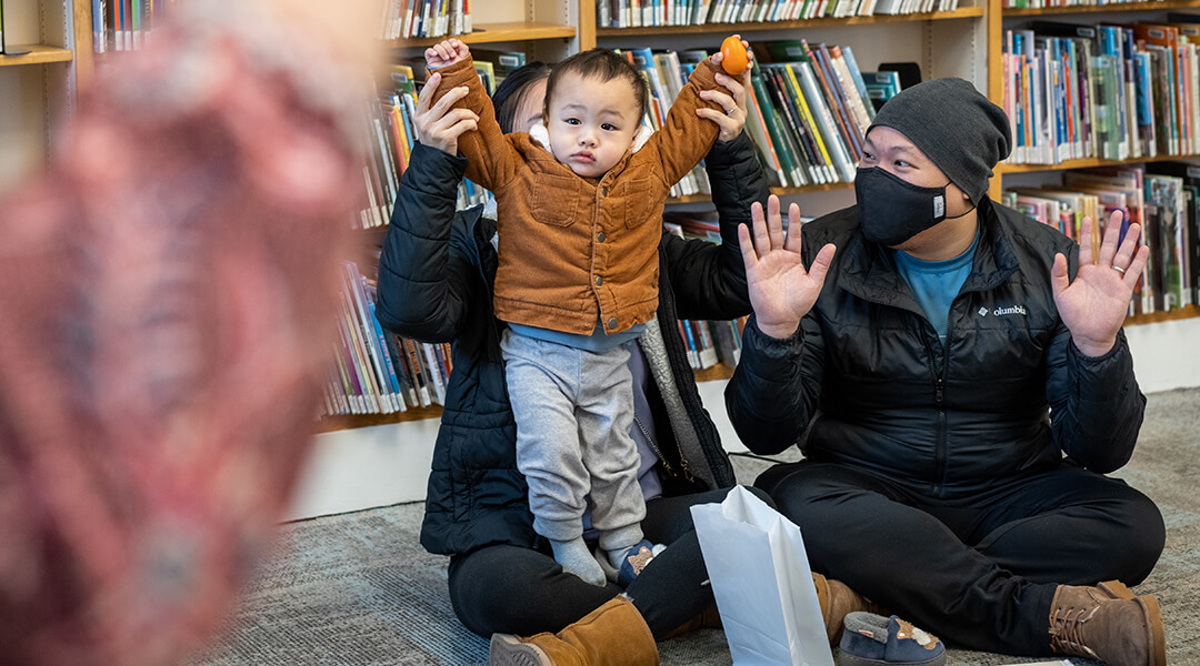 A caregiver helps a toddler stretch tall during a storytime rhyme.