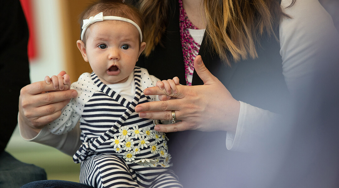 Close-up of an infant sitting on a caregivers lap, holding their fingers with their small hands.