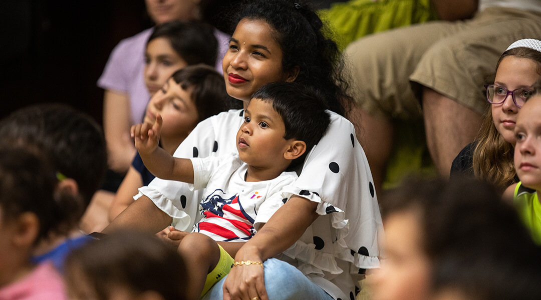 A young child, about two, sits on their caregivers lap during a crowded storytime.