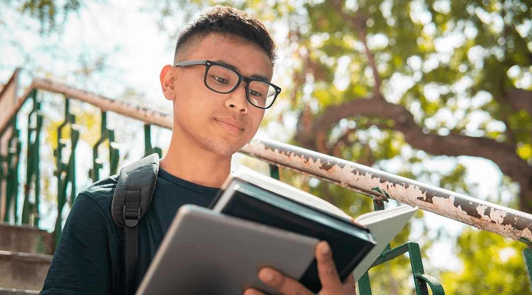 Teen boy wearing glasses and reading a book outside under a leafy tree