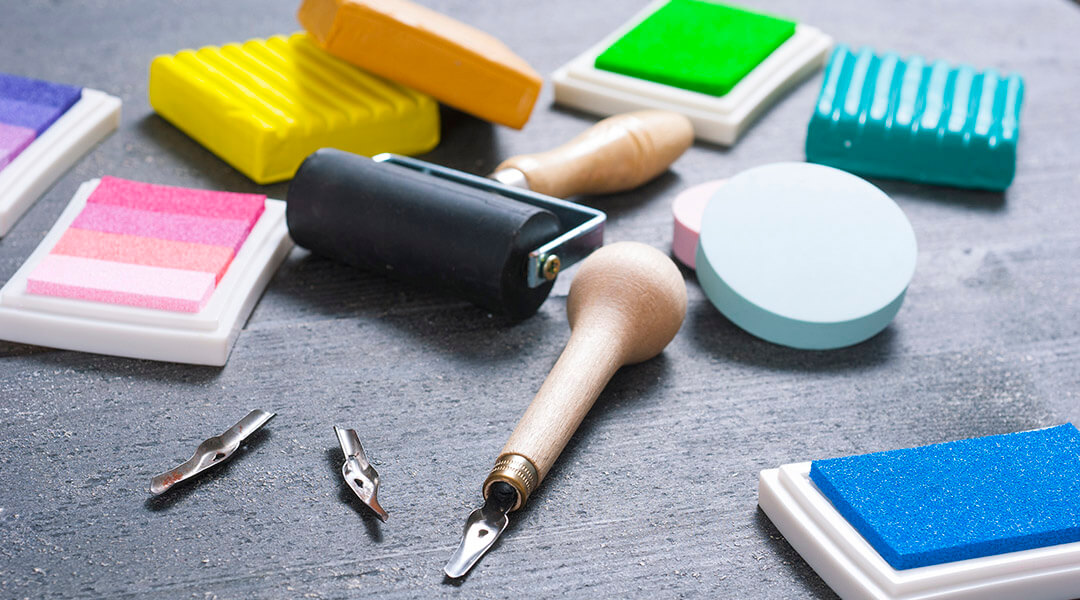 Printmaking tools on a tabletop including pads, plasticine blocks, brushes, paint roller and carving cutters.