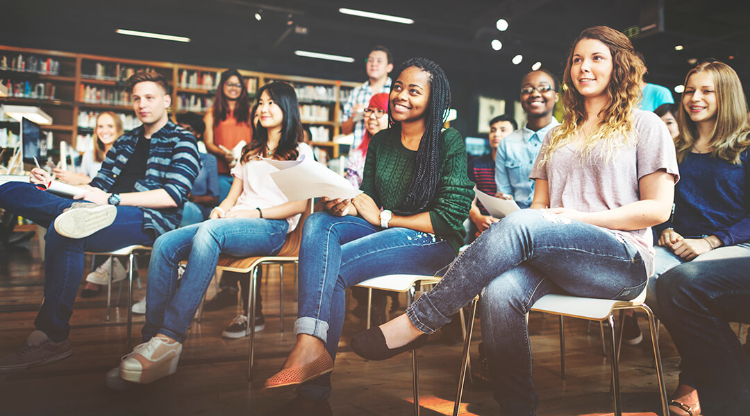 An audience of young adults listen to a presentation by an unseen speaker in a library.