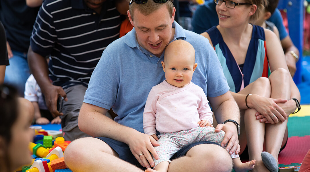 An infant sits on their caregivers lap during a playtime crowded with other children, adults, and toys in the library.