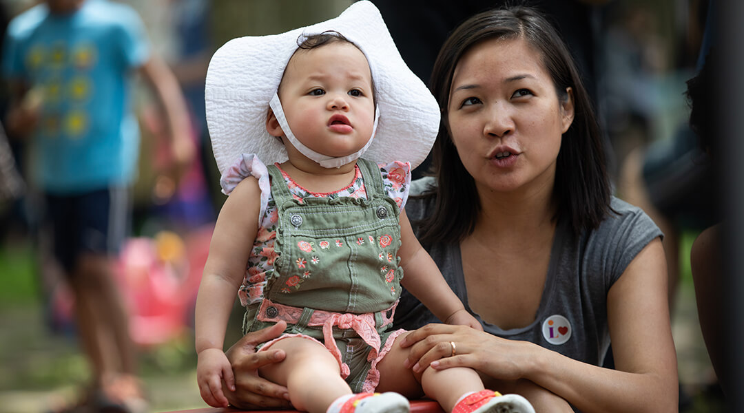 A caregiver sits with a baby in a sunhat during an outdoor storytime.