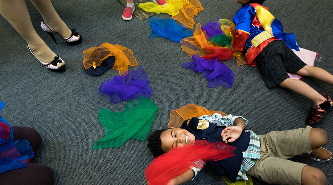 Two young children, about four wear costumes and play on the floor among scarves during storytime.