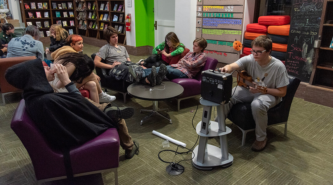 A group of teenagers lounging around the teenspace, one playing with a ukulele and small amp.