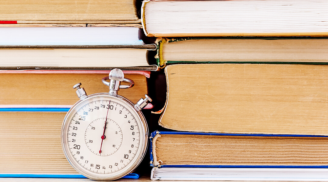Stack of many books and stopwatch on simple backdrop.