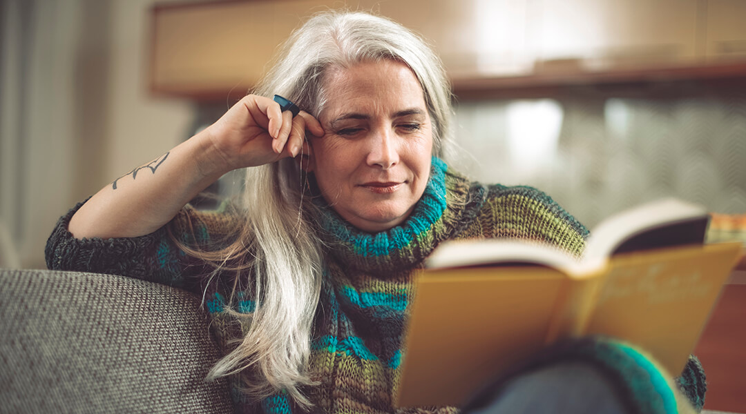 A mature adult with long white hair and a tattoo on their wrist reads in the living room.