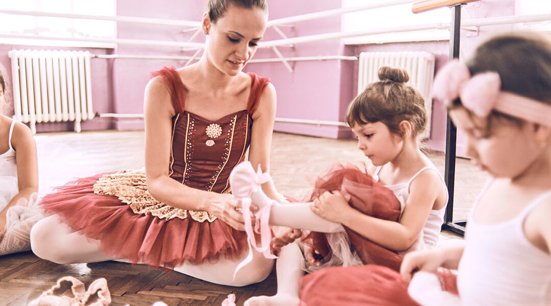 An adult in a dark rose tutu helps young dancers, around four, into their ballet flats in a dance studio.