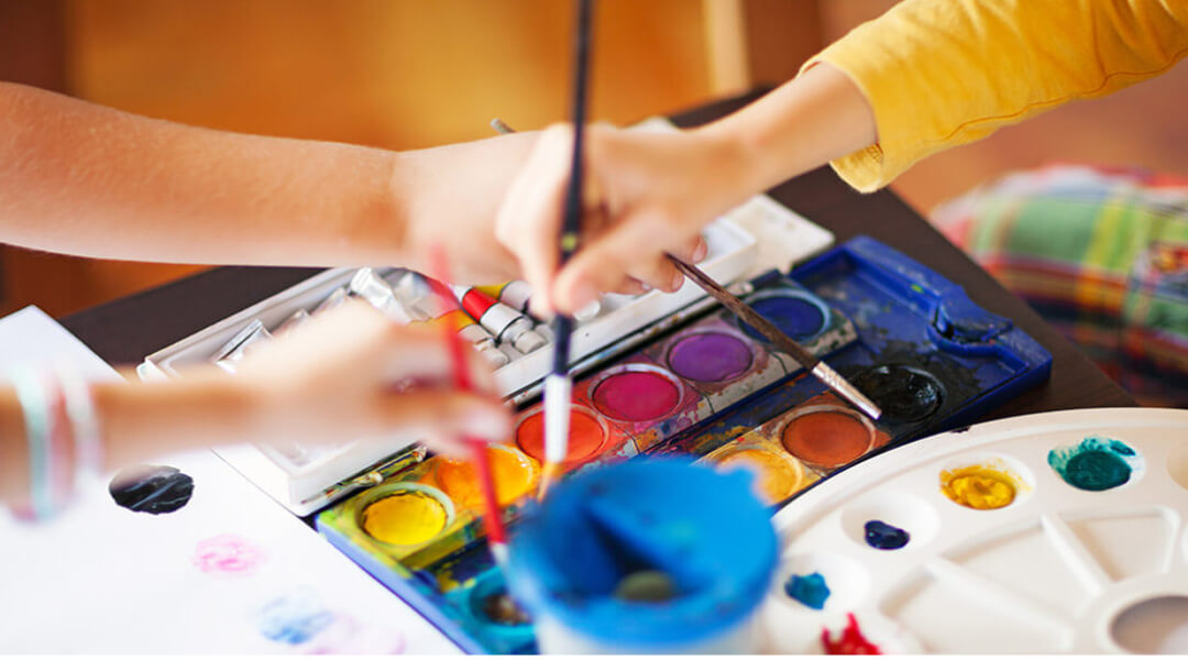 Close-cropped image of children reaching to share a palette of watercolor paints.