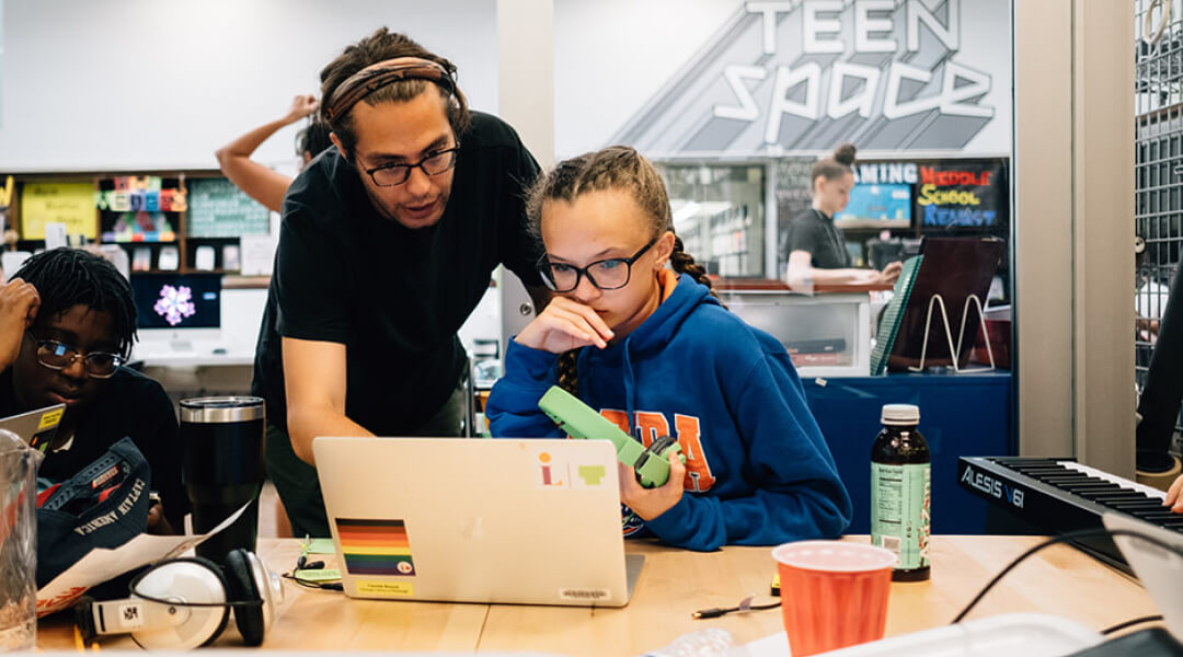 A librarian assists a teenager on a laptop in the library's teen space.