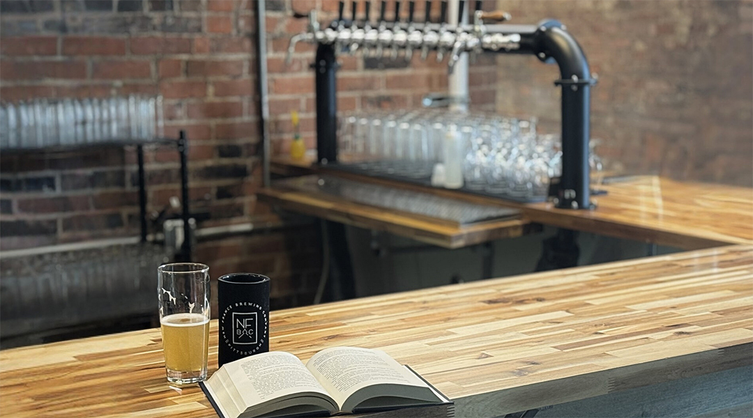 An open book, pint glass of beer, and travel mug with the New France Brewing Company logo on an empty bar; beer taps are in the background.