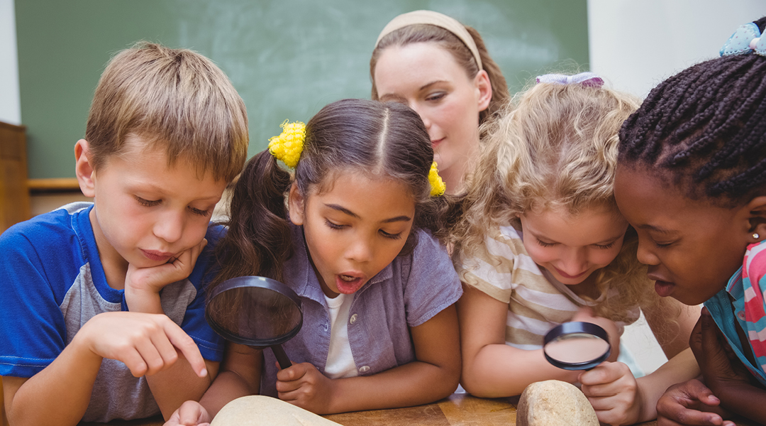 A small group of elementary school children crowd around a desk to inspect rocks with magnifying glasses.