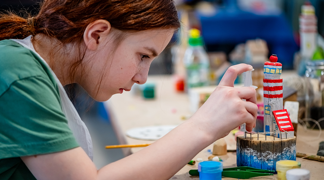 A child, about ten, works on painting a model lighthouse surrounded by craft supplies.