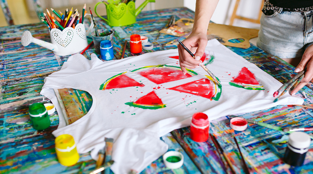 Hands apply paint to a white t-shirt, drawing slices of watermelon.