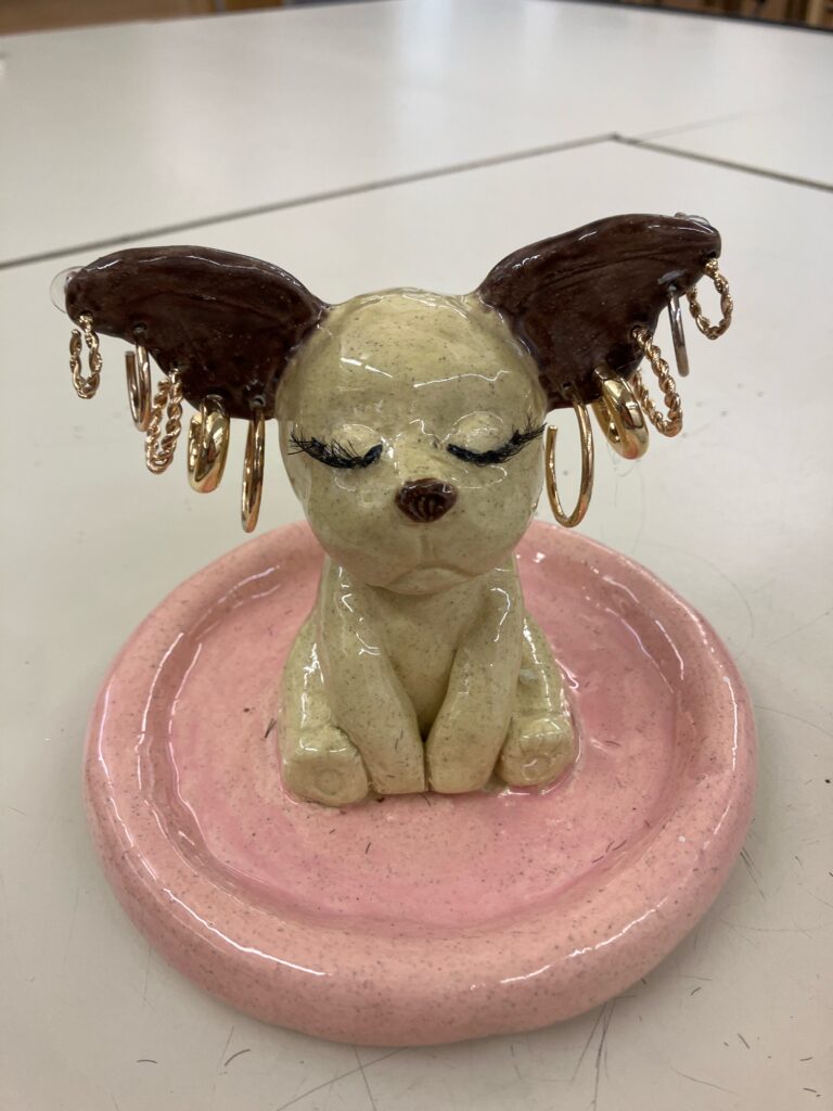 By Jessica Shullo. A small ceramic earring holder with a pale pink base featuring a dog sitting down - the ears hold five pairs of hoop earrings.