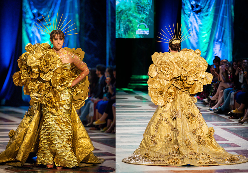 A runway model wearing a flowing gold dress with golden flower embellishments along the front and train.