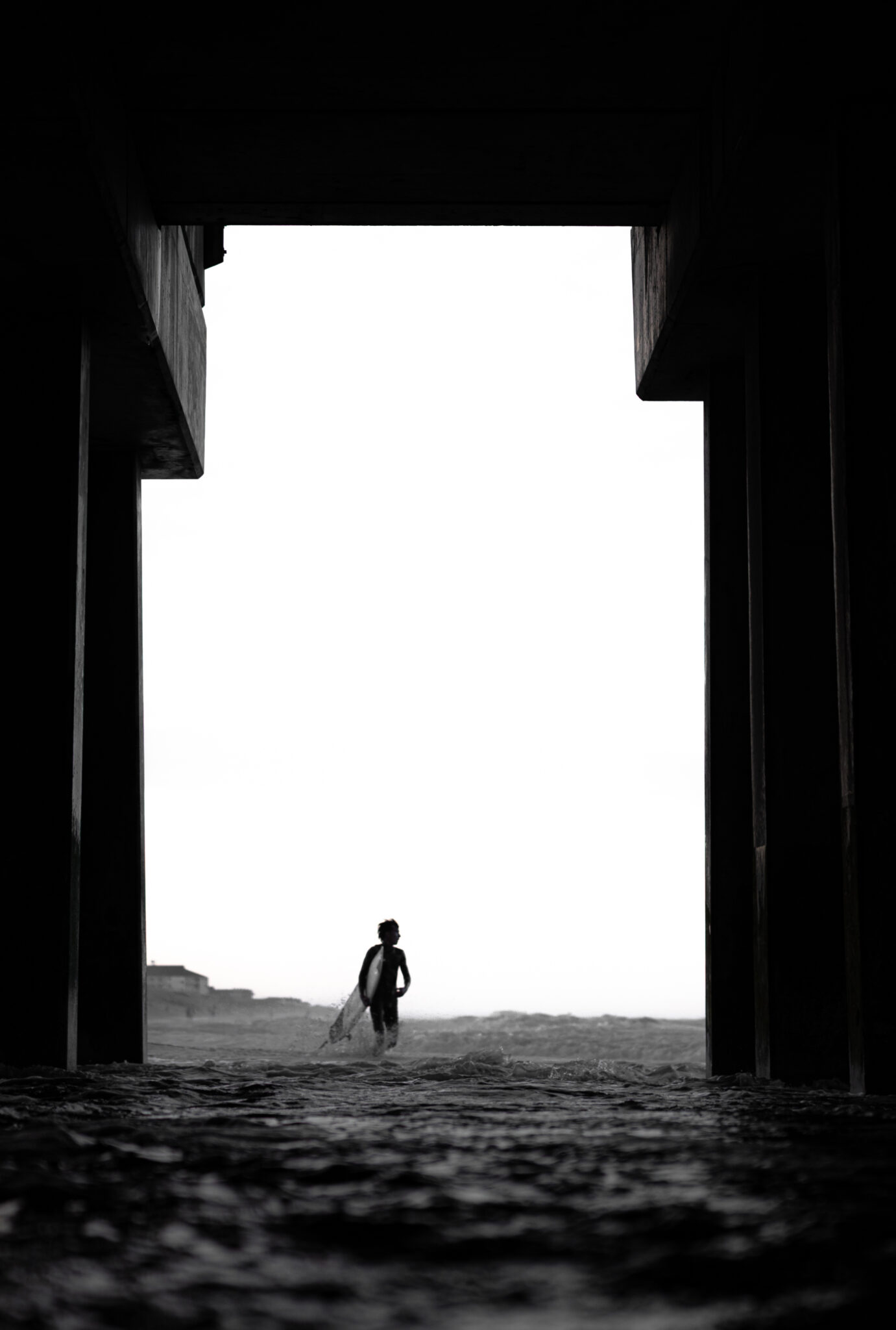 A surfer walks along the beach, framed by a concrete tunnel.