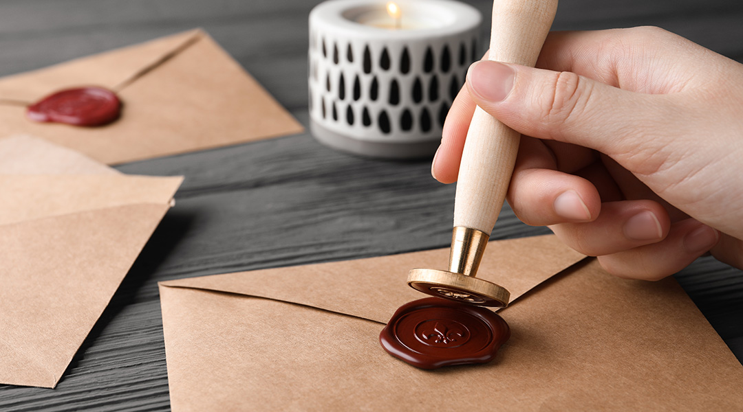 A hand presses a stamp into a wax seal on the back of a letter.