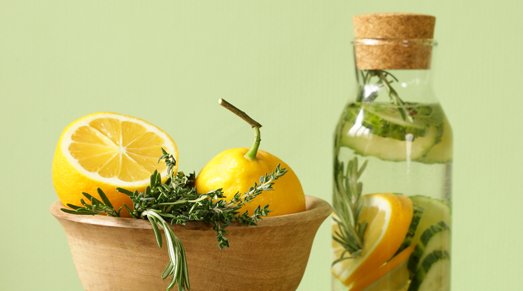 Lemons and herbs in a bowl next to a glass bottle filled with lemon, herbs, and water.