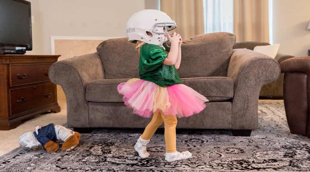 A toddler wearing a tutu and football helmet in a living room.