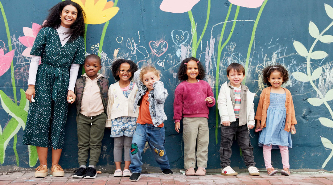 A teacher and six toddlers pose in front of a mural of flowers and chalk drawings.