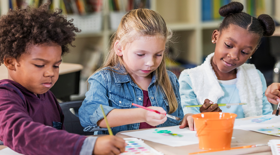 Three elementary-school aged children paint in a classroom.