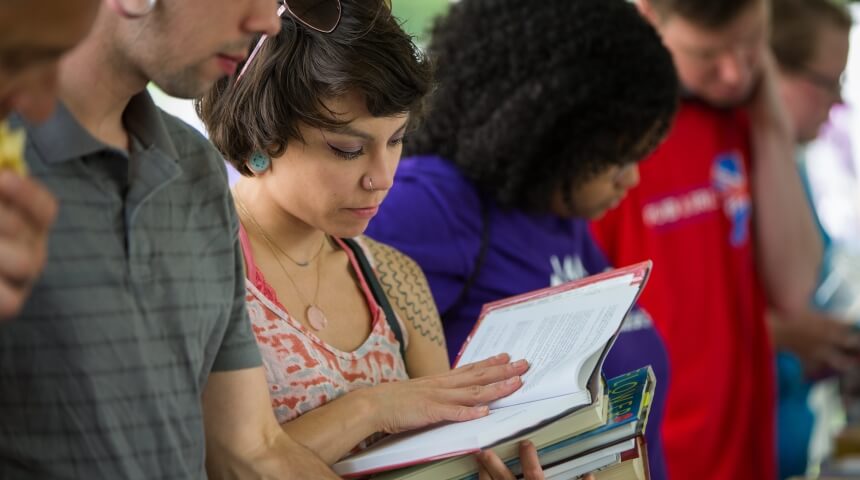 Young people stand in a line. One is holding a stack of books and reading the top book.