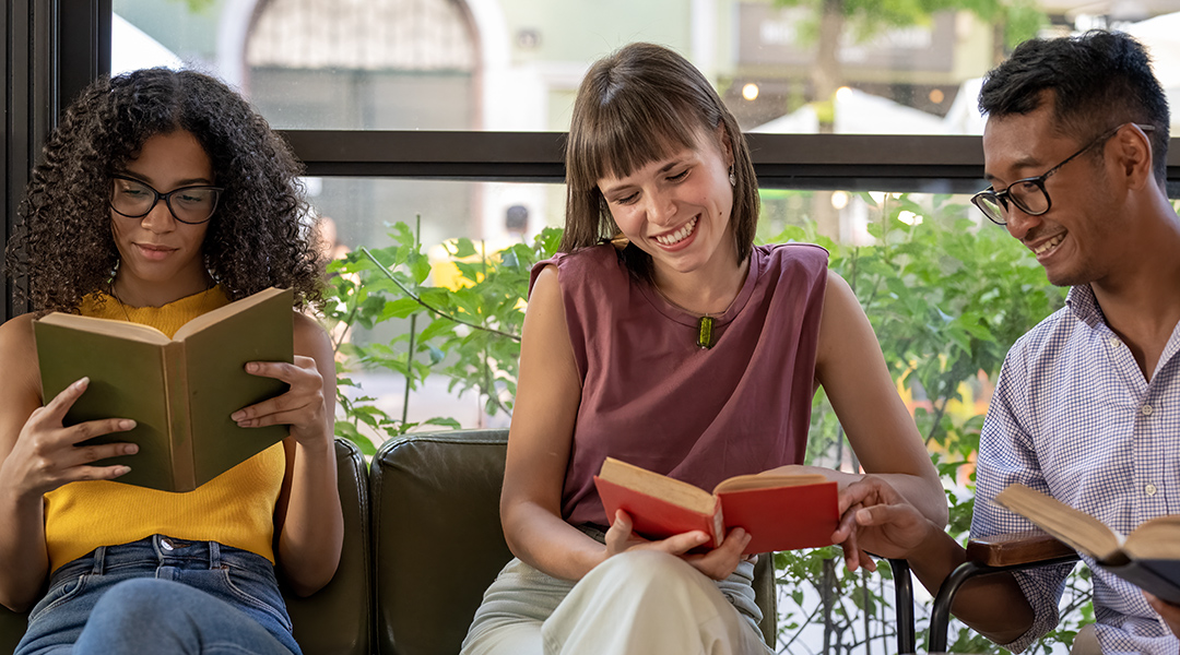 Three young adults sitting by a window all enjoying their own book.