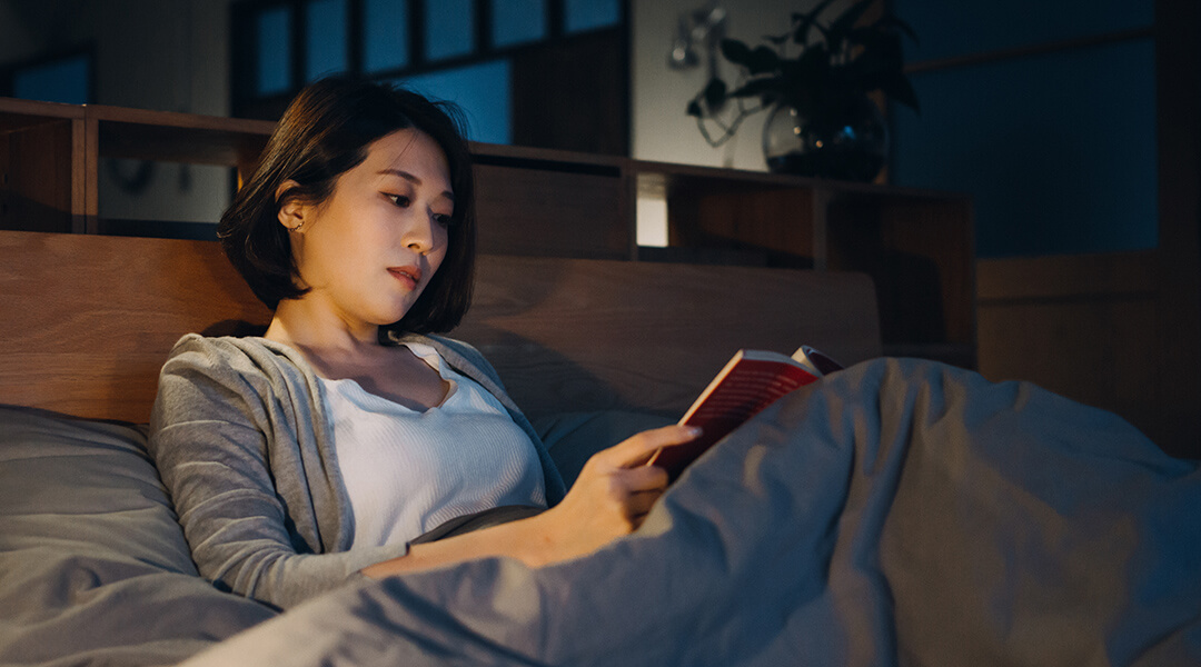 A woman relaxes in her bed with a book at night.