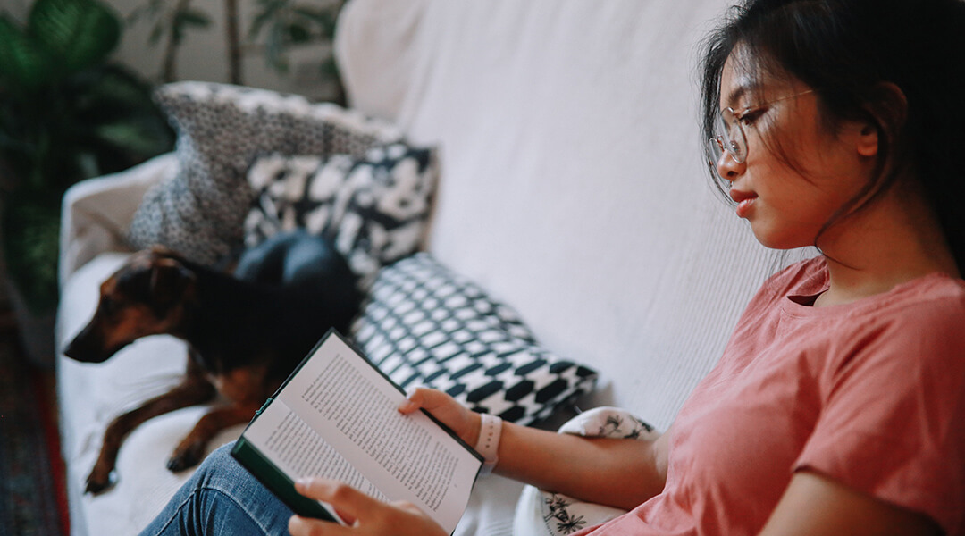 An adult reads on a couch next to a pet dog