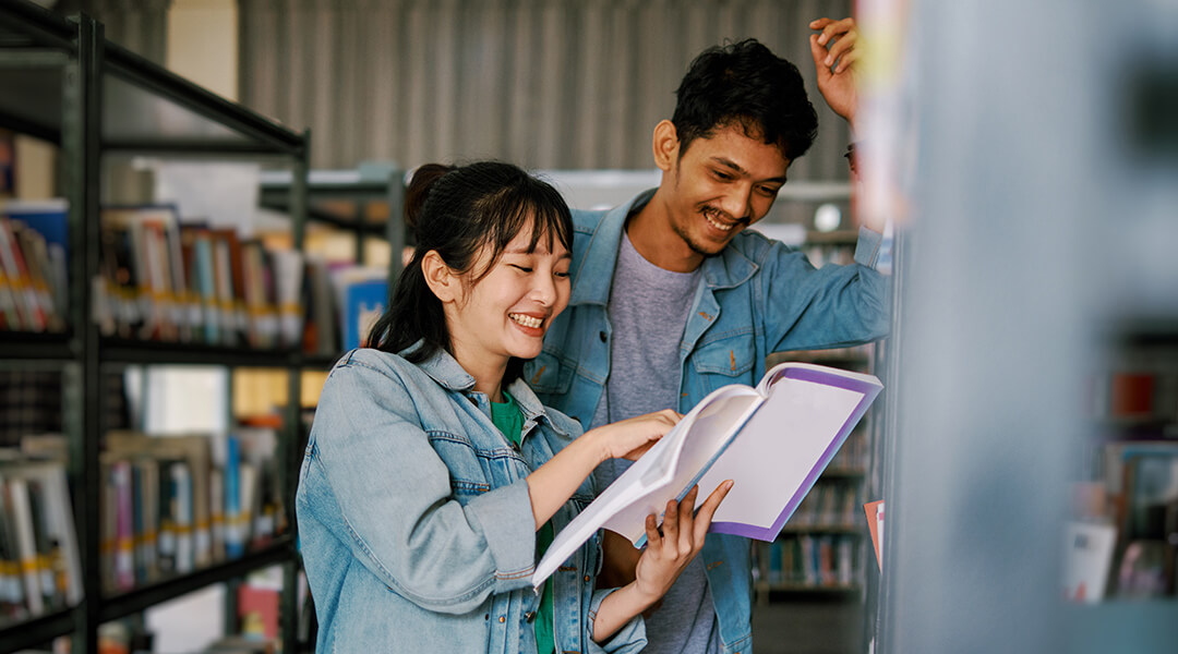 A woman and man laugh over a book in the middle of a library aisle