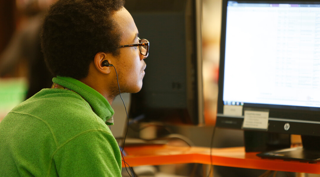 A man looks at a public computer screen while listening to earplugs that are connected to the computer.